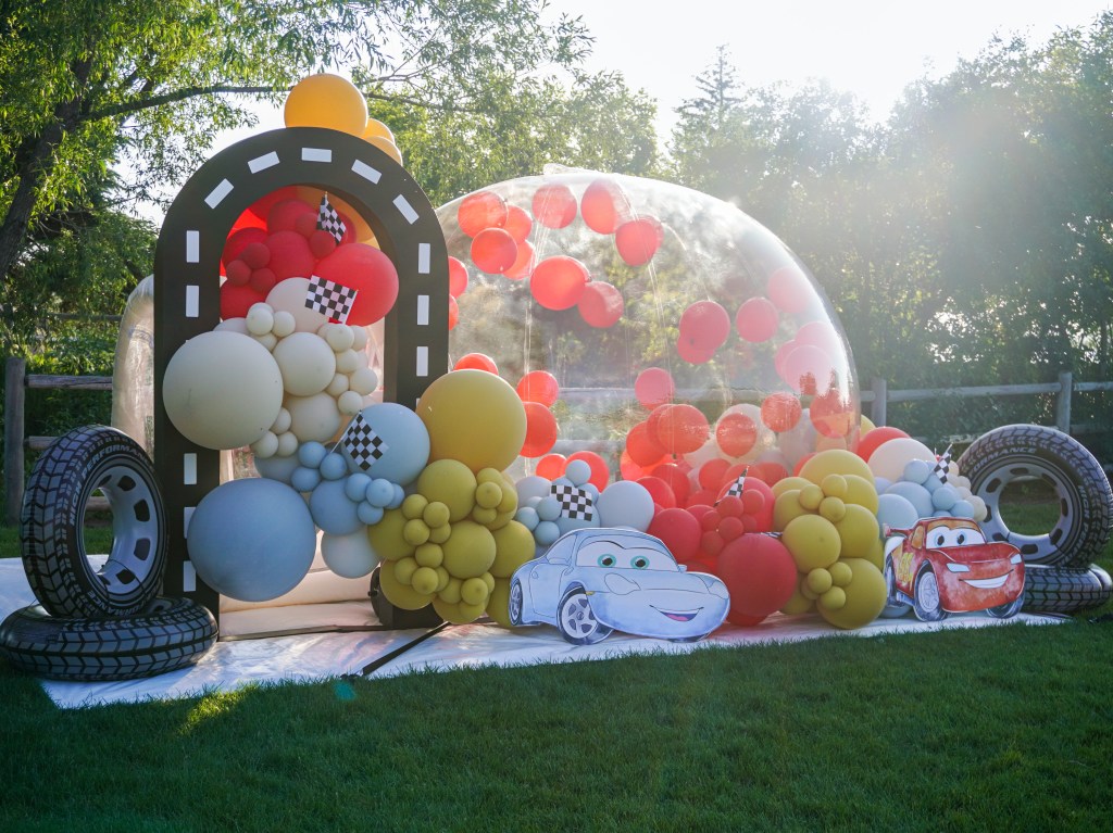 Luxury Cars-themed bubble house gender reveal setup in Denver by Chuchi's Events, featuring custom balloon garland, red and orange balloons, outdoor party rental decor, and life-size Lightning McQueen and Sally cutouts.