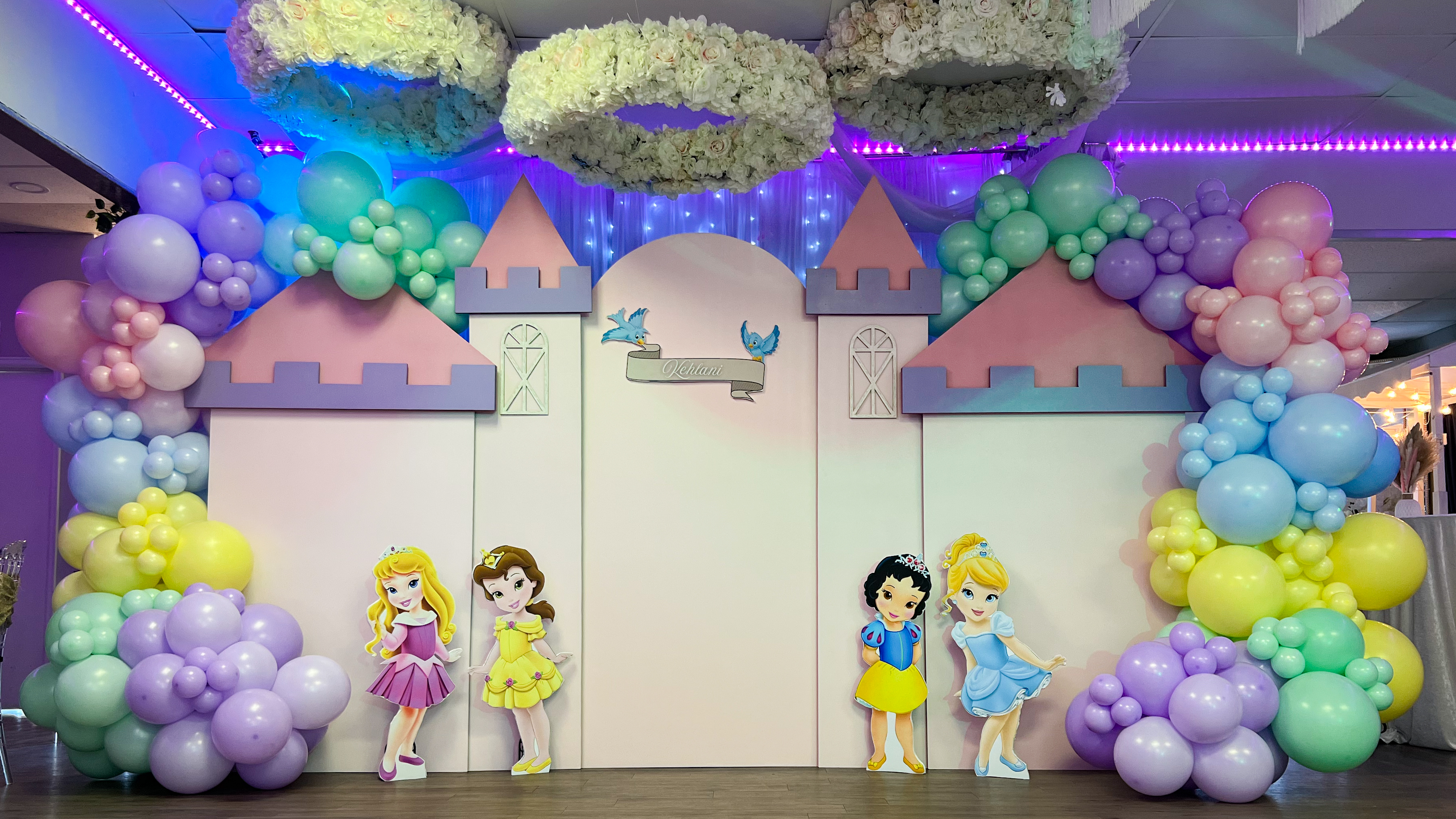 Whimsical princess-themed party backdrop featuring a pastel castle setup with custom name signage, surrounded by colorful balloon garlands in shades of pink, purple, blue, yellow, and green. Adorned with adorable princess cutouts, including Cinderella, Belle, Snow White, and Aurora, this luxury event decor creates a magical fairy-tale atmosphere. Elegant floral chandeliers and soft lighting enhance the enchanting design, making it the perfect setup for a princess birthday party or themed celebration in Denver.