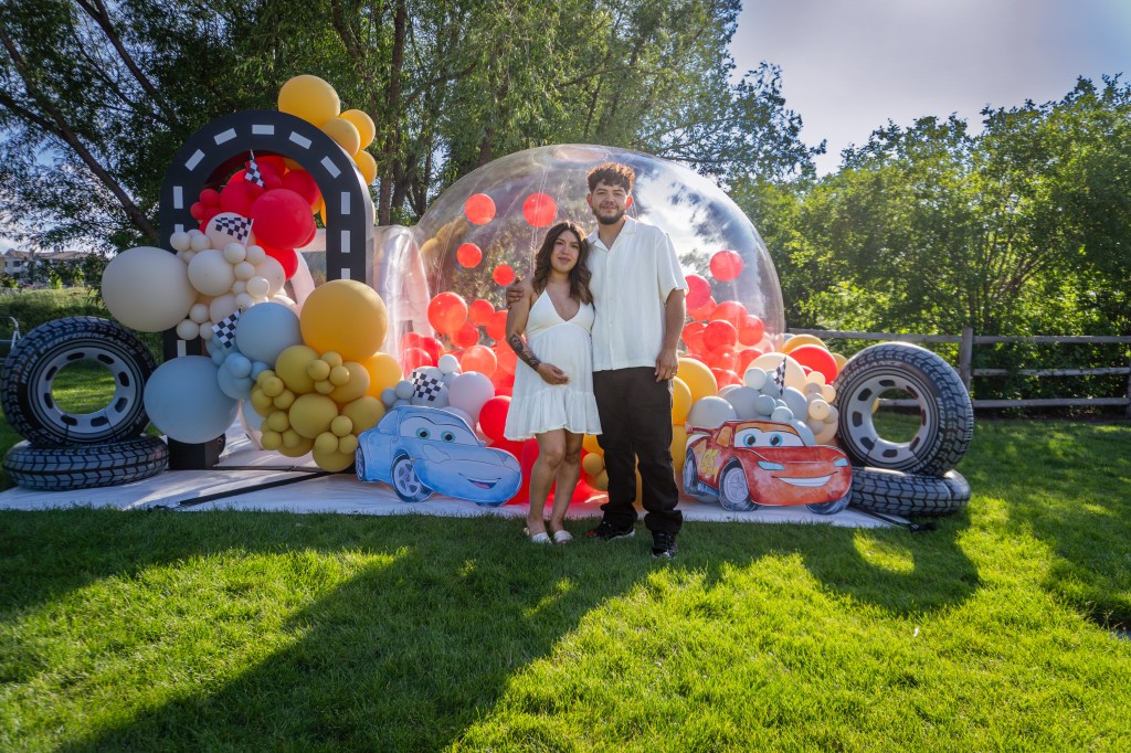 Luxury Cars-themed bubble house gender reveal setup in Denver by Chuchi's Events, featuring custom balloon garland, red and orange balloons, outdoor party rental decor, and life-size Lightning McQueen and Sally cutouts.