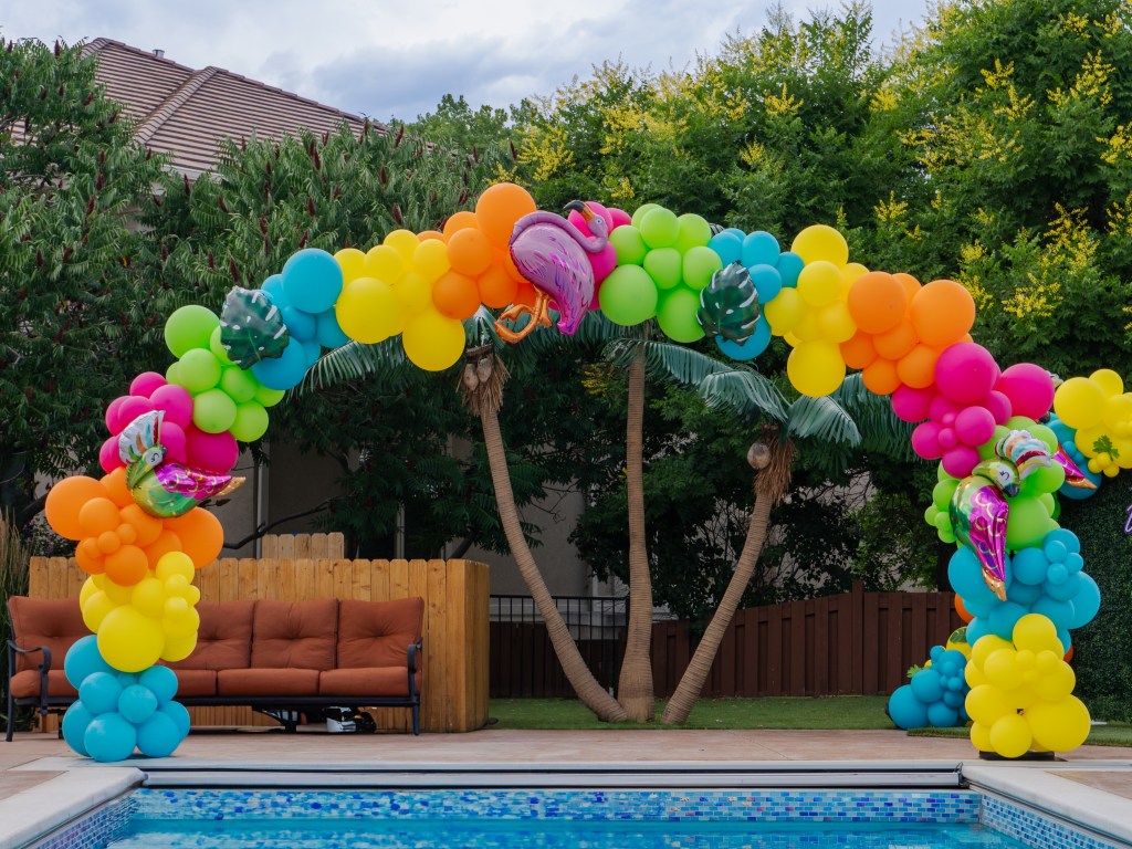 Tropical birthday party balloon arch setup by Denver event rental company featuring rainbow balloons, grass backdrop, and poolside decor for kids party