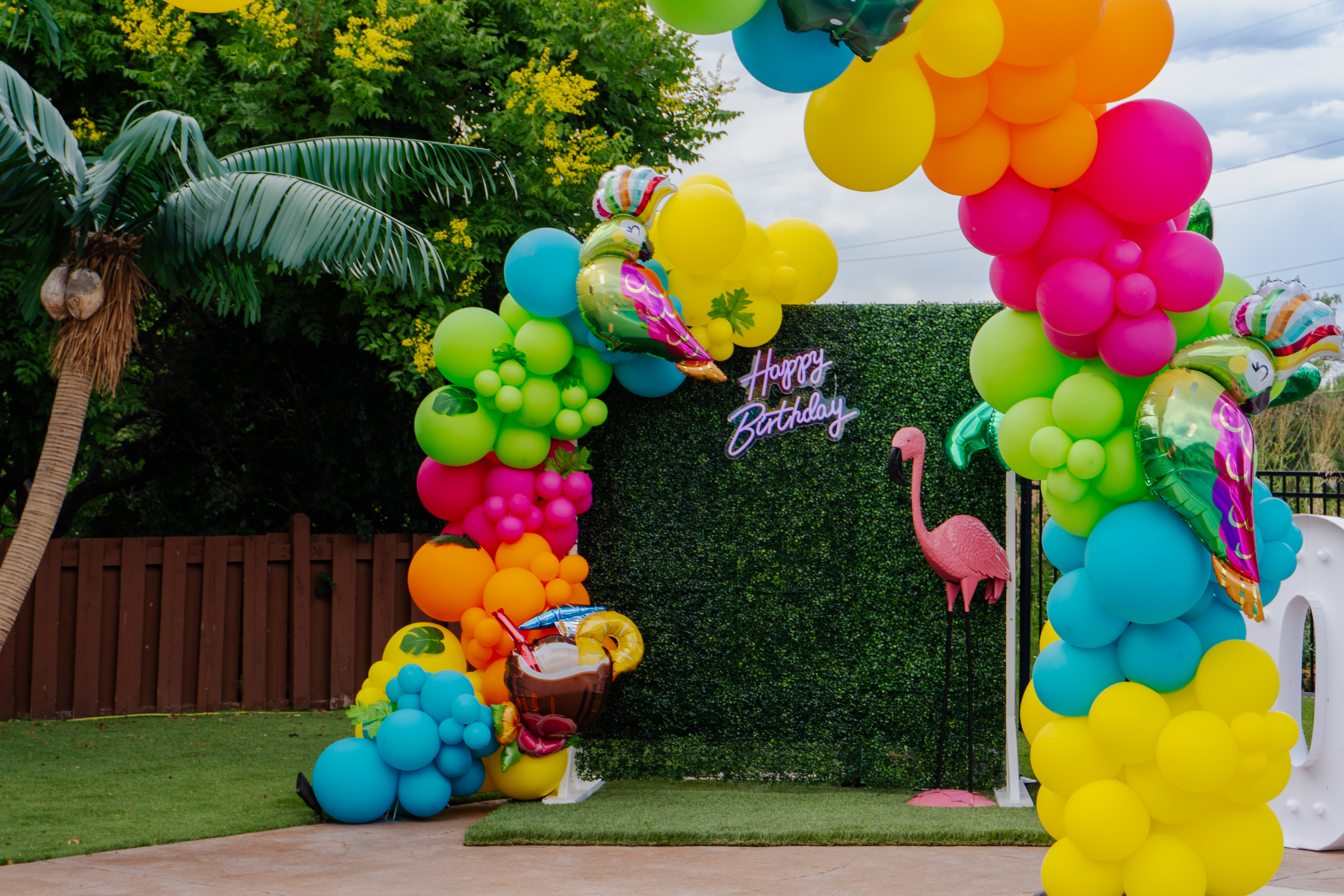 Tropical birthday party balloon arch with rainbow balloons and custom grass backdrop by Denver event rental company, perfect for kids party or summer celebration by the pool