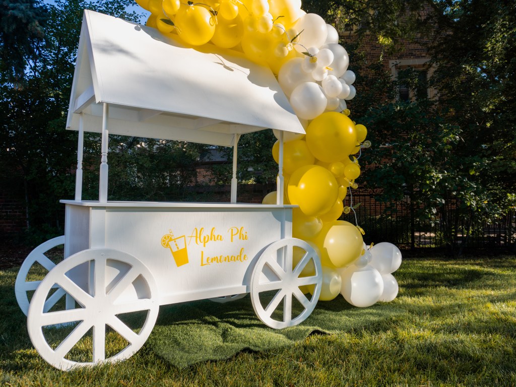 A charming white dessert cart rental decorated with a custom Alpha Phi lemonade logo and accented with a vibrant yellow and white balloon garland. Set up outdoors on a sunny day in Boulder, Colorado, this lemonade-themed cart is ideal for sorority events, graduation parties, and summer celebrations. Perfect for serving drinks, treats, or desserts, this stylish cart adds a festive and Instagram-worthy touch to any event.