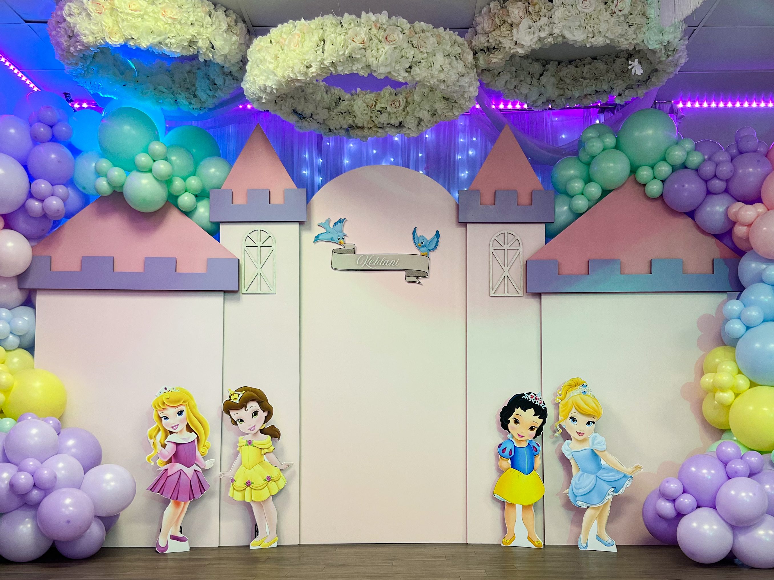 Whimsical princess-themed party backdrop featuring a pastel castle setup with custom name signage, surrounded by colorful balloon garlands in shades of pink, purple, blue, yellow, and green. Adorned with adorable princess cutouts, including Cinderella, Belle, Snow White, and Aurora, this luxury event decor creates a magical fairy-tale atmosphere. Elegant floral chandeliers and soft lighting enhance the enchanting design, making it the perfect setup for a princess birthday party or themed celebration in Denver.