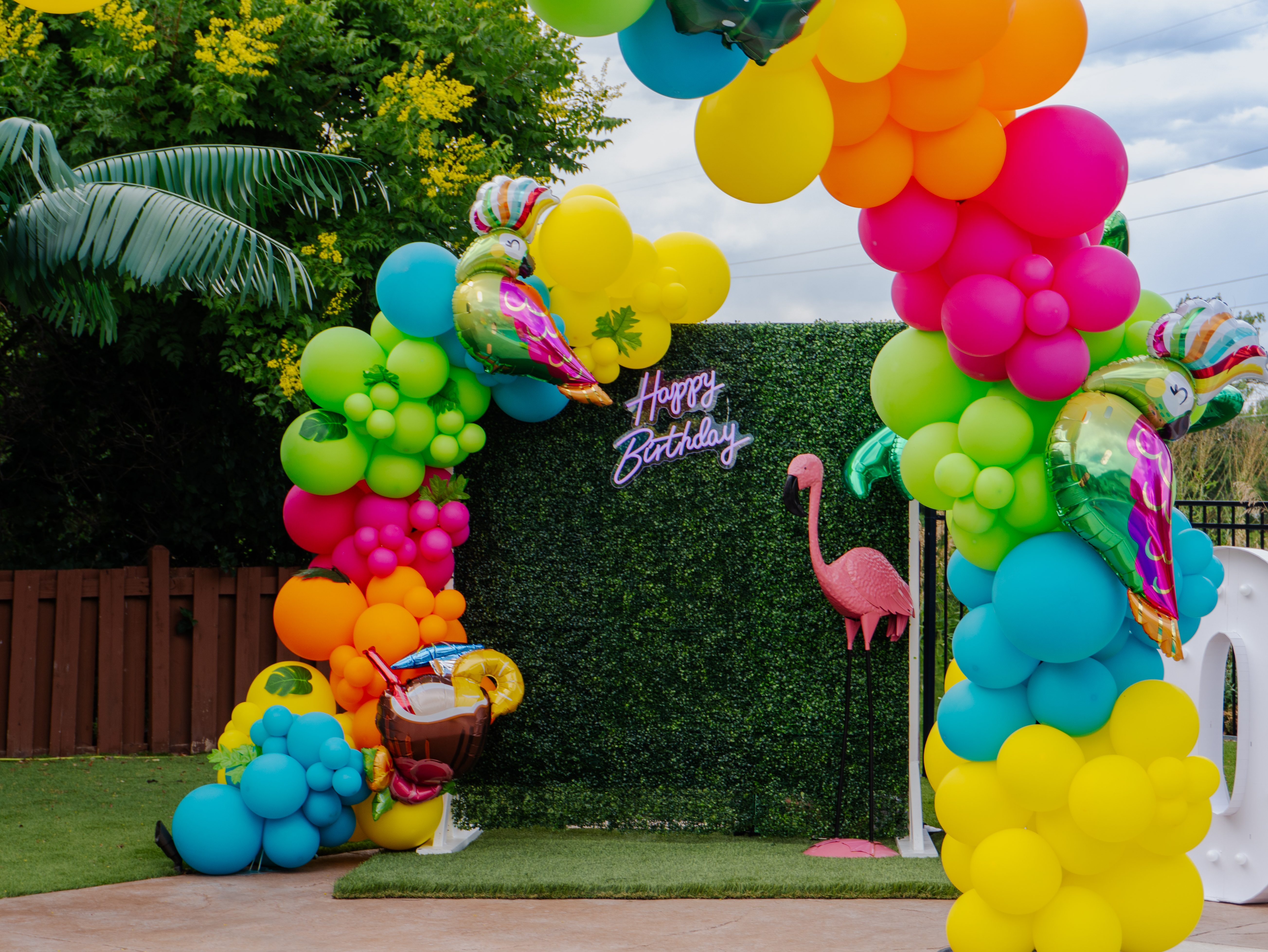 Tropical birthday party balloon arch with rainbow balloons and custom grass backdrop by Denver event rental company, perfect for kids party or summer celebration by the pool